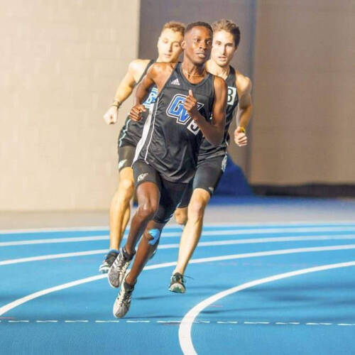 Three male runners compete on an indoor blue track, with the foreground athlete wearing a black singlet marked “GV L” sprinting around the curve. Two competitors trail close behind as the lead runner pushes forward with focused determination.